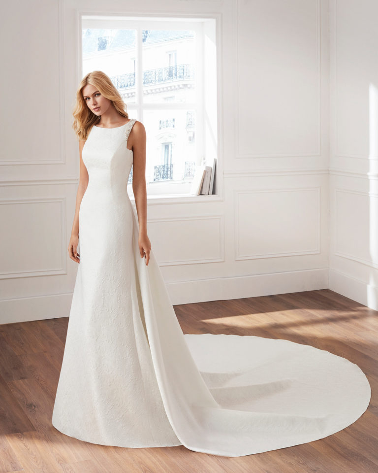 Vestido de novia estilo clásico en brocado. Escote barco, detalles de pedrería y cola desmontable. Disponible en color natural. Colección  2019.