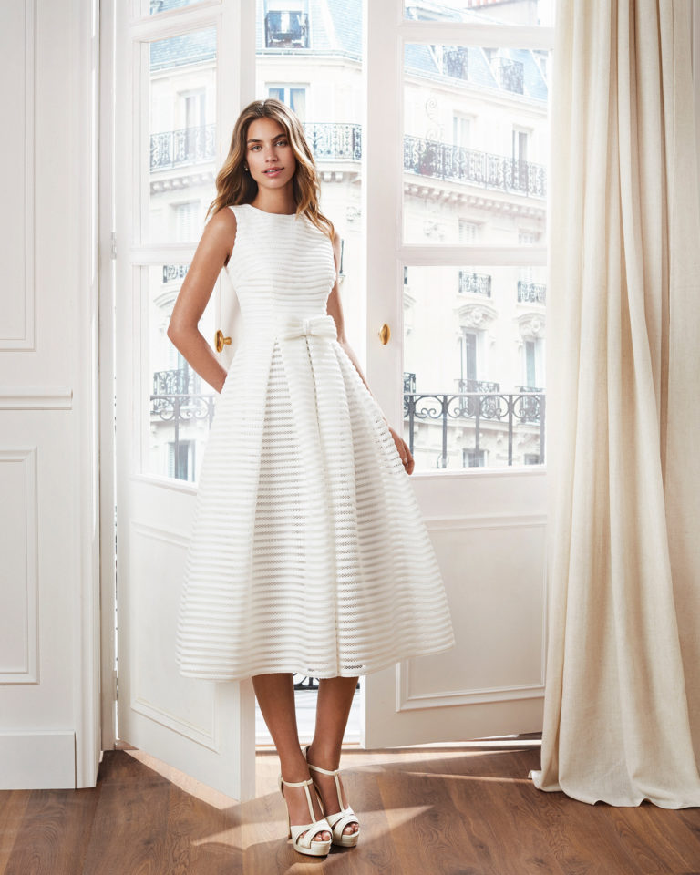 Vestido de novia corto estilo clásico. Escote barco, espalda en V y pliegues en falda. Disponible en color natural. Colección  2019.