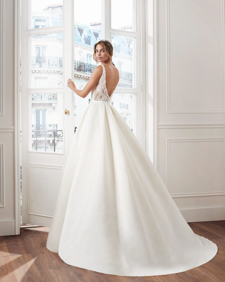Vestido de novia estilo clásico línea A en raso. Escote en V, cuerpo de encaje pedrería y pliegues en cintura. Disponible en color natural. Colección  2019.
