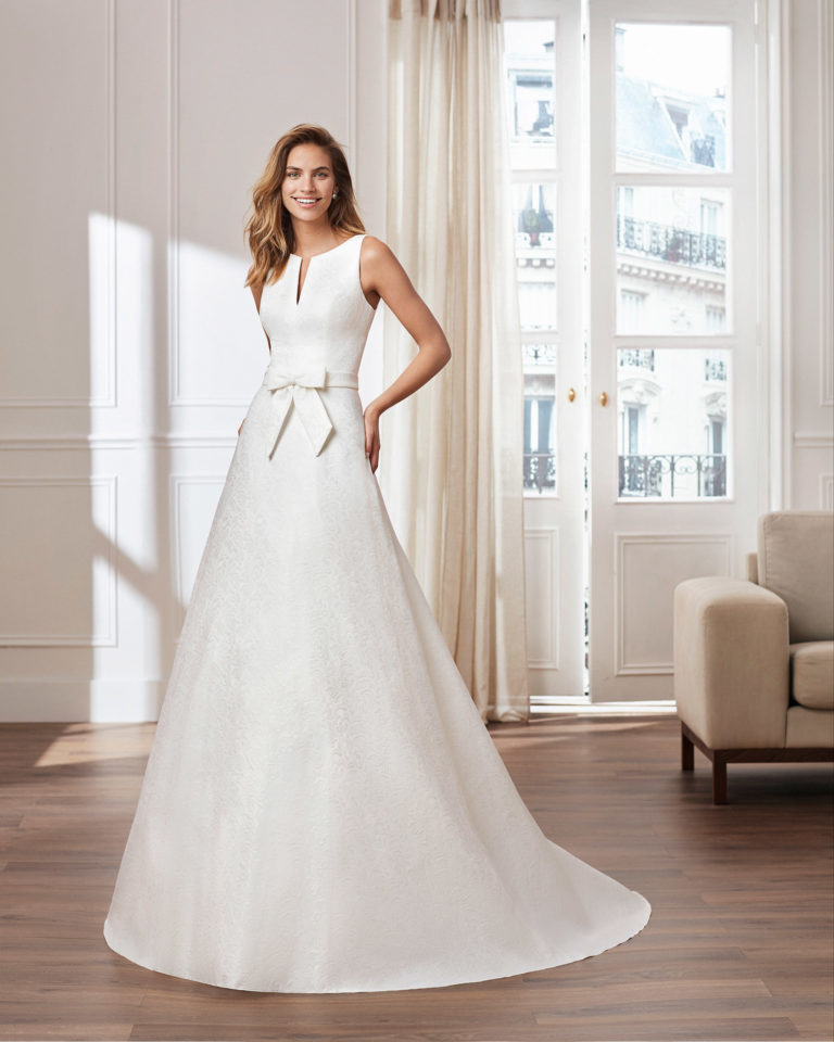 Vestido de novia estilo clásico línea A en brocado. Escote en V en cuerpo y en espalda. Disponible en color natural. Colección 2020.