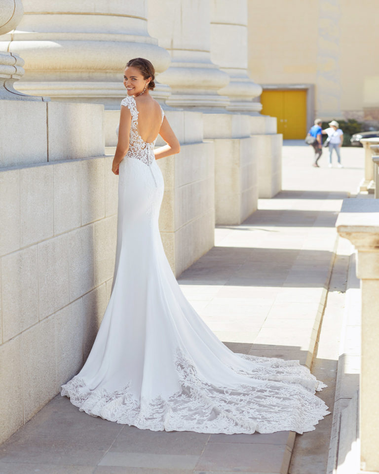 Sheath-style wedding dress in stretch crepe and lace. Bateau neckline with sweetheart underlay, low back, short sleeves with appliqués and trefoil train. 2021  Collection.