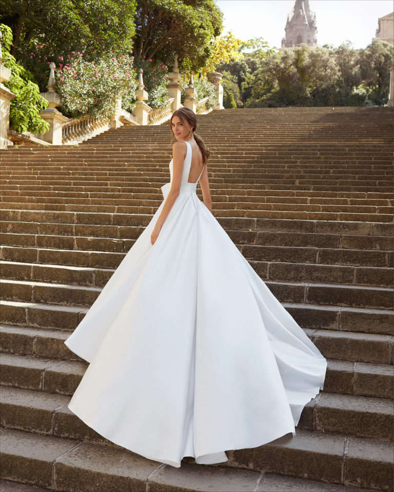 Classic-style wedding dress in satin. Deep-plunge neckline and U-back, with front bow at waist and pockets. 2021  Collection.