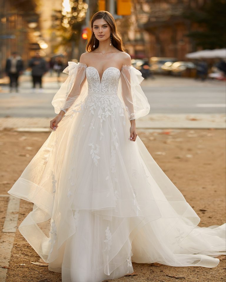 Romantic princess-style wedding dress, made of tulle and lace appliqués, with a sheath-style skirt with flounces; with a deep-plunge neckline with delicate beadwork detail, an open back, and voluminous tulle sleeves. Unique Luna Novias outfit made of tulle with lace appliqués. LUNA_NOVIAS.