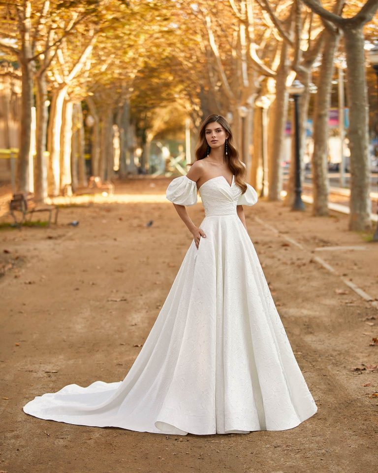 Classic-style wedding dress, made of fiore brocade, featuring a full buttoned skirt; with a strapless neckline, a button-up back, and puffed sleeves. Luna Novias look made of fiore brocade. LUNA_NOVIAS.