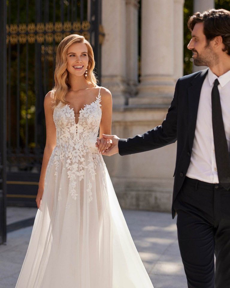Vestido de novia largo de estilo romántico, confeccionado en tul, encaje y pedrería. Se completa con un escote en V, una espalda escotada, tirantes y una flor de adorno. Un modelo de tendencia Luna Novias. LUNA_NOVIAS.