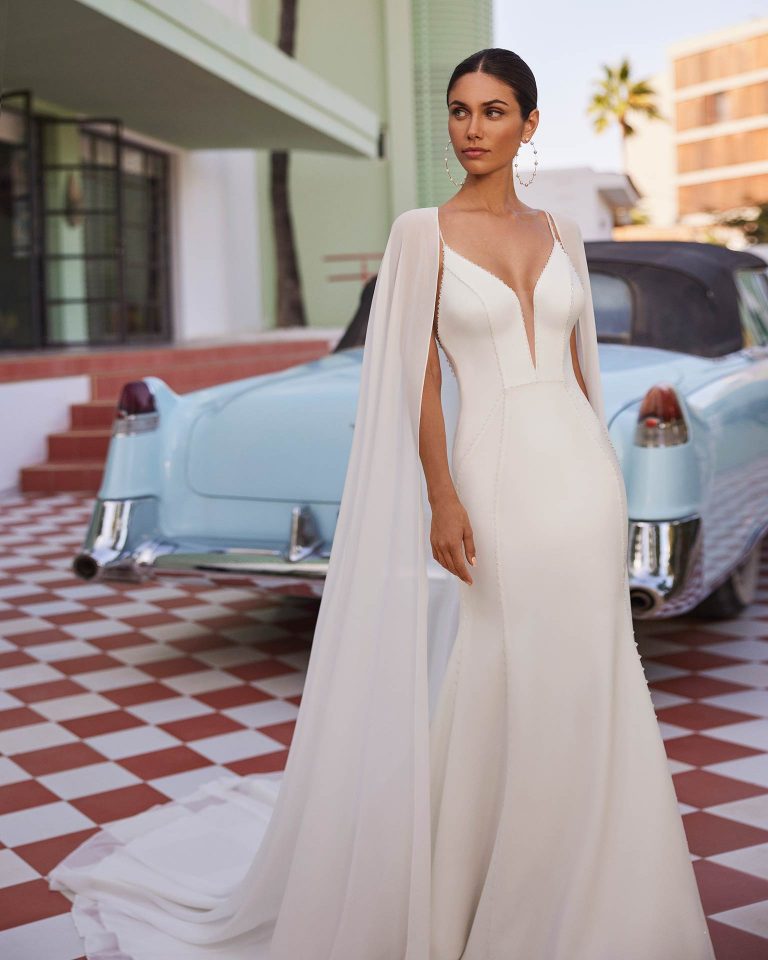 Vestido de novia largo de dos piezas con corte sirena. Crepé bielástico Vera, escote pico, espalda con tirantes cruzados y capa. LUNA_NOVIAS.