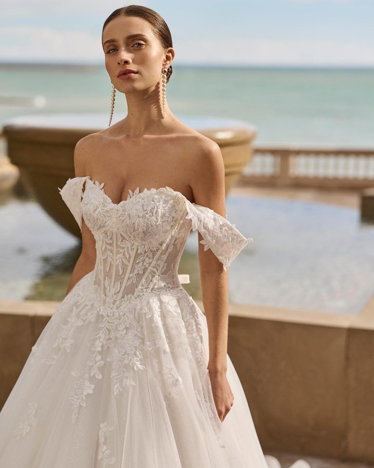 A-line wedding dress with sweetheart neckline, off-shoulder straps and corset closure. Lace bodice with beading, tulle skirt with appliqués. LUNA_NOVIAS.