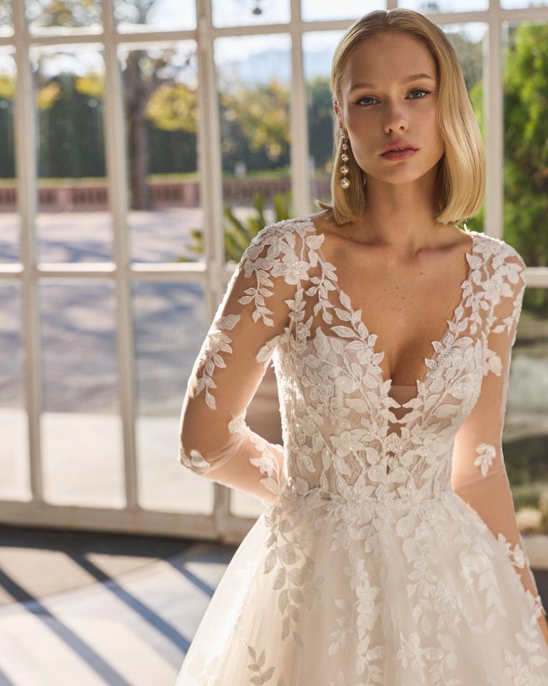 A-line wedding dress with deep neckline and long sleeves. Lined lace bodice, illusion back. Tulle skirt. Appliqués on bodice, sleeves and skirt. LUNA_NOVIAS.
