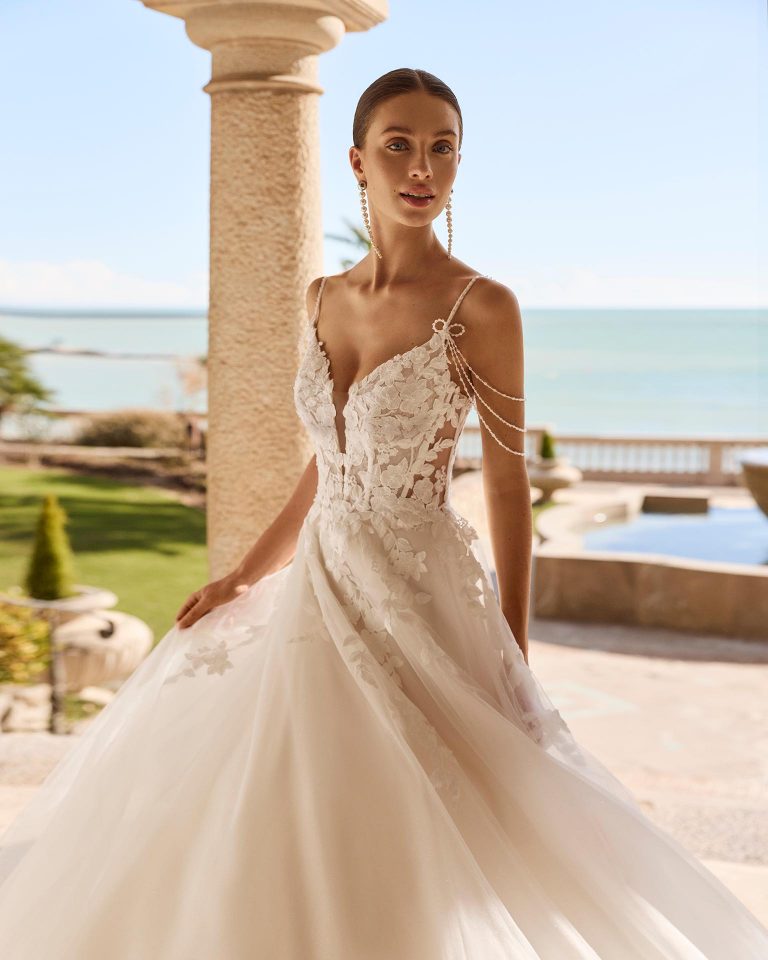 A-line wedding dress with deep neckline, straps and open back. Lace bodice with beadwork, glitter tulle skirt with appliqués. LUNA_NOVIAS.
